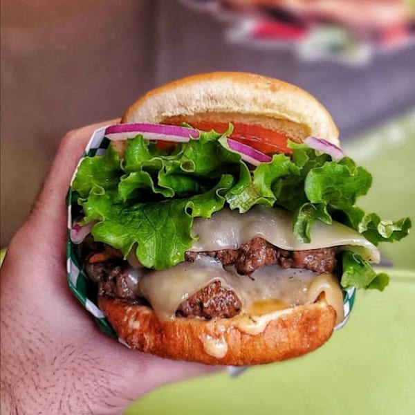 Hand holding a Burganic Hub double cheeseburger with lettuce, tomato, and red onion.